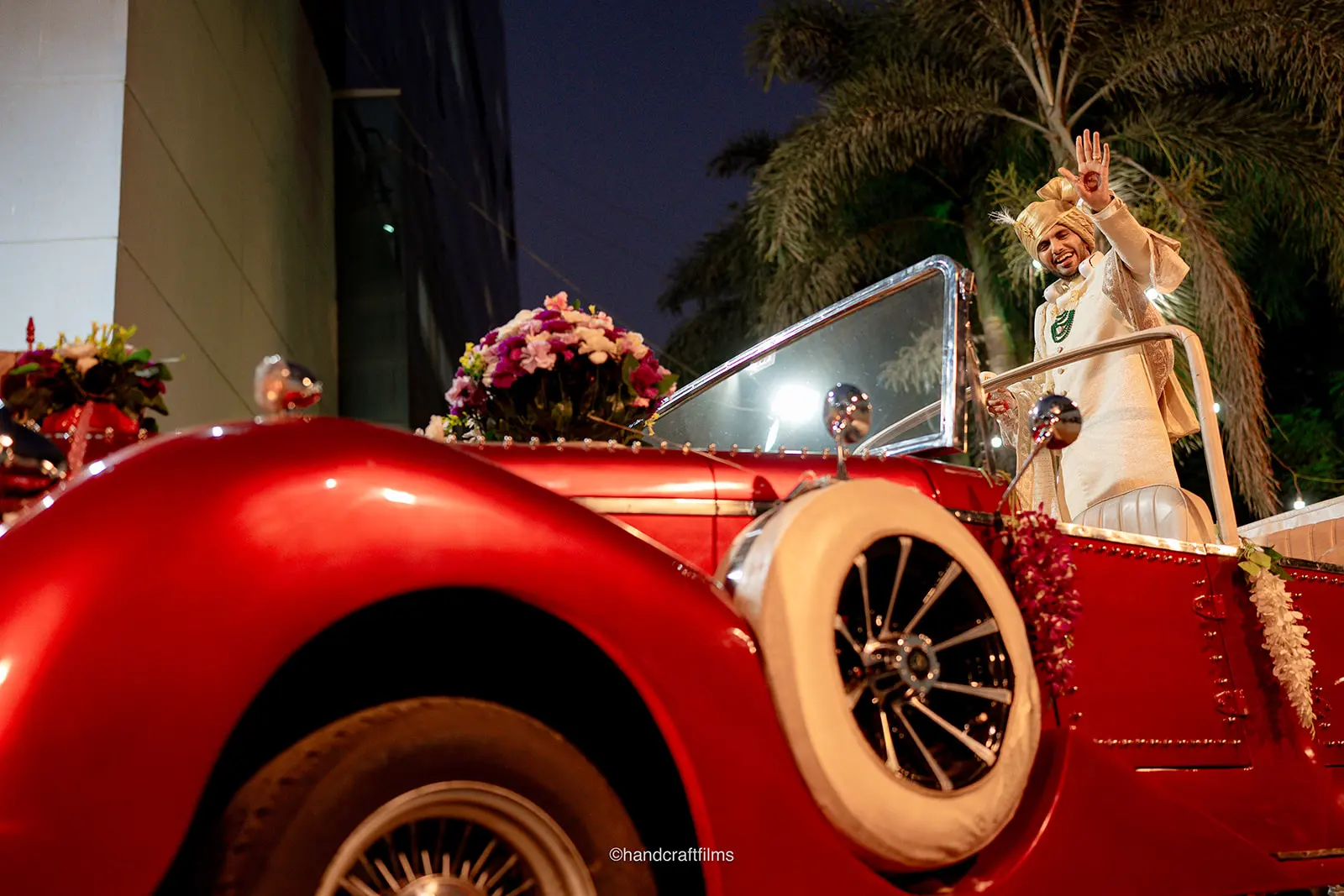 Groom waving from a vintage car during his evening Baraat, decorated with flowers